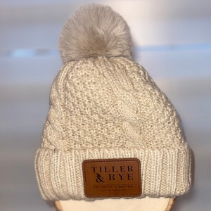 Puede incluir: Gorro de punto trenzado color crema con un pompón de piel sintética. El gorro tiene un parche de cuero marrón con el texto "Tiller & Rye Brewer Maine". Un gorro de invierno.