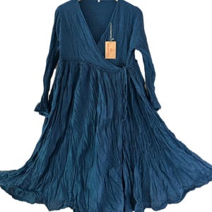 May include: A long-sleeved, dark teal wrap dress featuring a V-neckline, a gathered waistline, and a full, flowing skirt with a crinkled texture. A small rectangular tan paper tag is attached to the neckline with a thin string.
