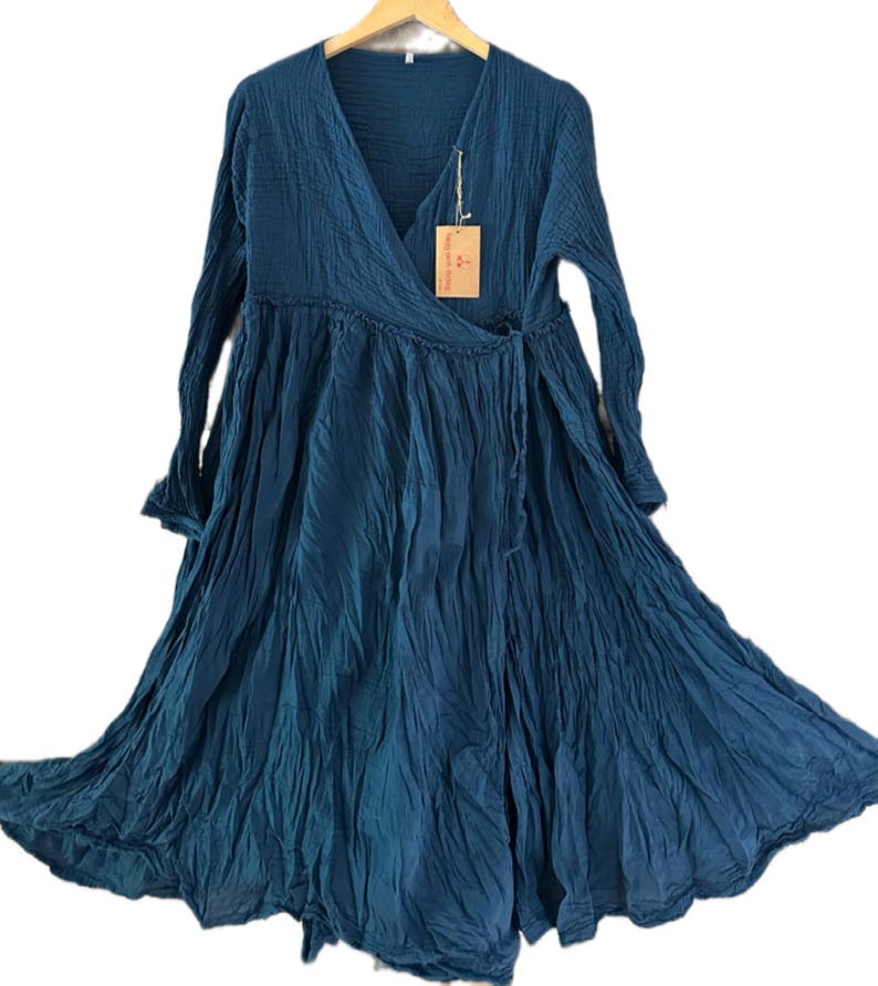 May include: A long-sleeved, dark teal wrap dress featuring a V-neckline, a gathered waistline, and a full, flowing skirt with a crinkled texture. A small rectangular tan paper tag is attached to the neckline with a thin string.