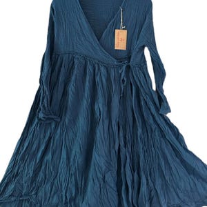 May include: A deep teal long-sleeved dress featuring a V-neckline, a wrap-style bodice with a side tie closure, and a gathered waistline. The fabric has a crinkled texture and includes a small rectangular brown paper tag attached to the neckline.