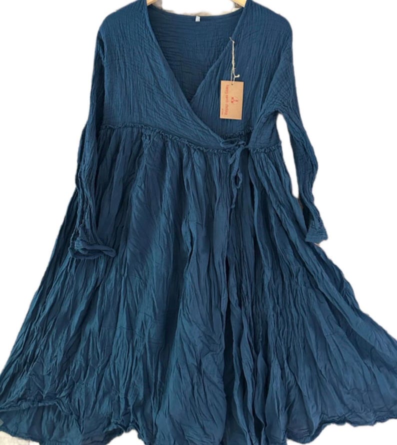 May include: A deep teal long-sleeved dress featuring a V-neckline, a wrap-style bodice with a side tie closure, and a gathered waistline. The fabric has a crinkled texture and includes a small rectangular brown paper tag attached to the neckline.