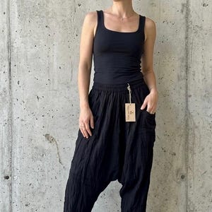 May include: A woman wearing a black tank top and black harem pants. The pants have large pockets and a loose, flowing fit. A small tag hangs from the pants. The background is a plain concrete wall.