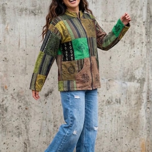May include: A long-sleeve zip-up jacket featuring a patchwork design in shades of green, brown, and black with various printed patterns including suns and geometric shapes, paired with light blue distressed wide-leg denim jeans.