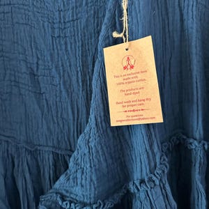 May include: A deep blue garment with a crinkled texture and a tiered design. A tan rectangular tag is attached with twine, featuring the text: This is an exclusive item made with 100% organic cotton. The products are hand-dyed. Hand wash and hang dry for proper care. For questions zeegeecollections@yahoo.com.