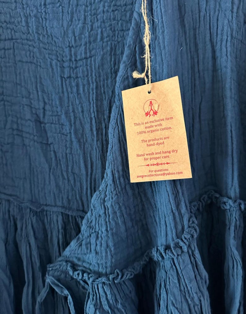 May include: A deep blue garment with a crinkled texture and a tiered design. A tan rectangular tag is attached with twine, featuring the text: This is an exclusive item made with 100% organic cotton. The products are hand-dyed. Hand wash and hang dry for proper care. For questions zeegeecollections@yahoo.com.