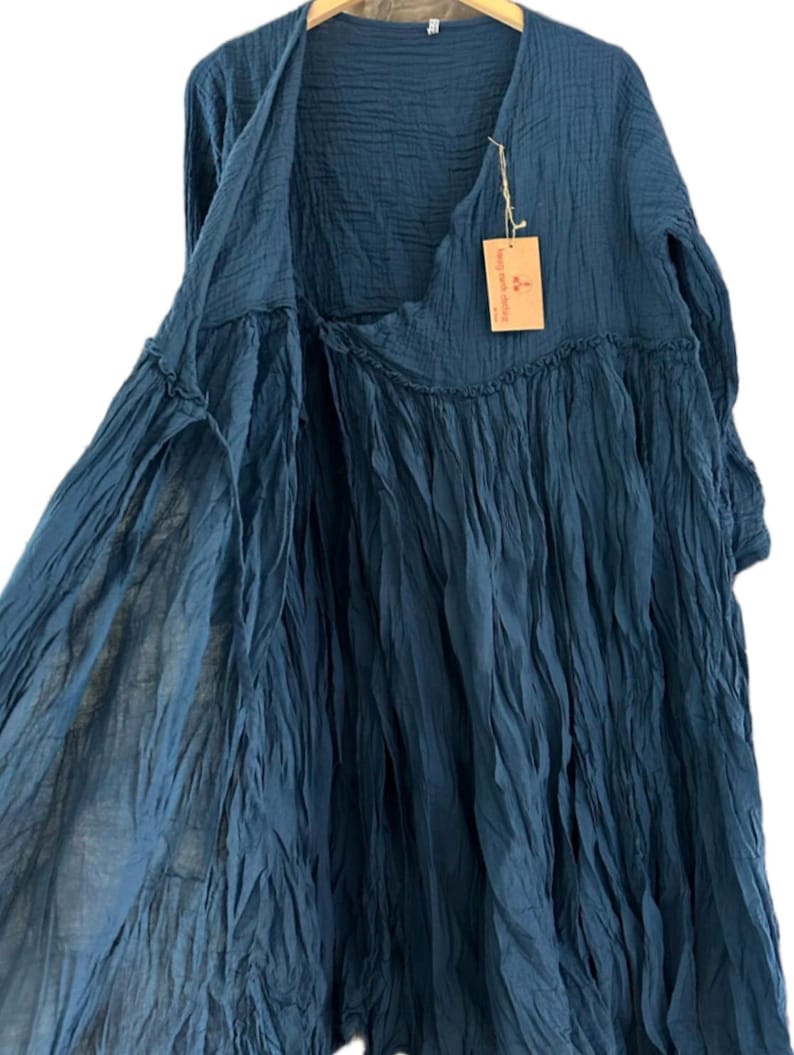 May include: A dark blue long-sleeved dress made of crinkled fabric with a wrap-style front, a gathered waistline, and a V-neckline. A small rectangular tan paper tag is attached to the neckline with string, featuring printed text and a logo.