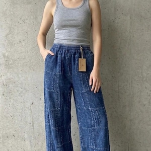 May include: A woman wearing a gray tank top and blue wide-leg pants. The pants have a patchwork design. The woman is also wearing sandals. The background is a plain gray wall.