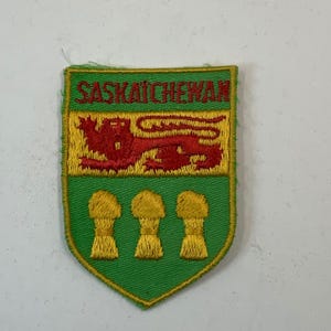 May include: Embroidered patch featuring the Saskatchewan provincial coat of arms. The shield-shaped patch has a green border and background. The design includes a red lion, a yellow upper section, and three golden wheat sheaves. The word "SASKATCHEWAN" is embroidered in red.
