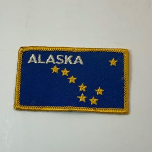 May include: Embroidered patch featuring the word "ALASKA" in white above a constellation of gold stars on a blue background. The patch has a gold border.