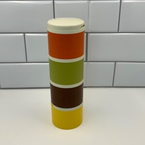 May include: A cylindrical, stackable storage container featuring four distinct colored sections separated by white rings. The top section is orange, followed by a lime green section, a dark brown section, and a bright yellow base section.