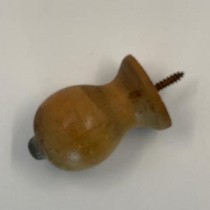 May include: A light brown wooden cabinet knob with a rounded, bulbous shape and a flared base. A metal screw protrudes from the back of the knob, and a small, circular silver-toned metal cap is visible on the front center of the wooden surface.