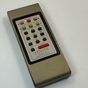 May include: A vintage Zenith Computer Space Command remote control. The remote is gray with a silver panel featuring black number buttons, and red and brown function buttons. The text "ZENITH COMPUTER SPACE COMMAND" is printed on the panel.