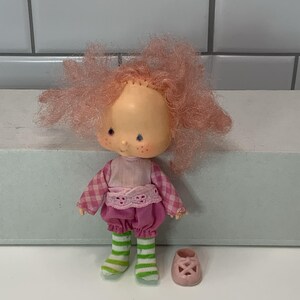 May include: A vintage doll with pink hair, freckles, and a light complexion. The doll wears a pink and white checkered shirt, pink shorts, and green and white striped socks. A pair of pink shoes with cut-out designs sits nearby.