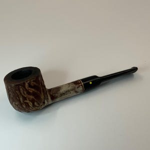 May include: A vintage tobacco pipe with a dark brown bowl and a light brown stem. The bowl has a textured design, and the stem is marked with the word "WILLARD". The mouthpiece is black with a yellow dot.