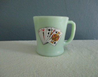 May include: A light green glass mug with a handle. The mug features a graphic of playing cards, including the King of Spades. The mug is likely vintage and suitable for coffee or tea.