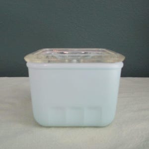 May include: A square, pale blue glass container with a clear, square lid. The container has a slightly frosted appearance and sits on a white surface. The background is a muted teal colour.