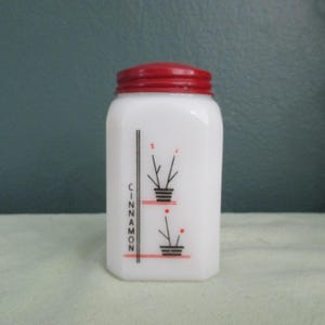May include: A white, rectangular spice jar with a red lid. The jar features a black and red floral design with the word "CINNAMON" printed vertically. A vintage kitchenware item.
