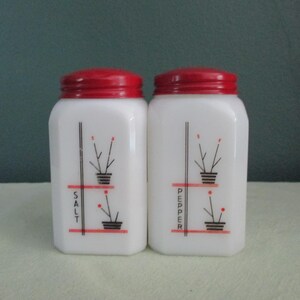 May include: A pair of vintage white salt and pepper shakers with red lids. Each shaker has a black and red floral design with the words "SALT" and "PEPPER" in black lettering.