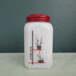 May include: A white glass nutmeg spice jar with a red lid. The jar has a black and red design with the word "NUTMEG" in black vertical text. The design includes two stylized plant illustrations.