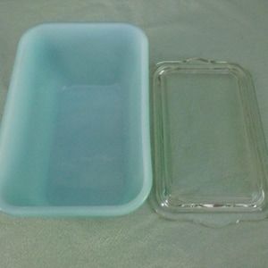 Op de afbeelding: Een vintage, lichtblauwe rechthoekige botervloot met een helder glazen deksel. De schaal heeft afgeronde hoeken en een licht taps toelopende vorm. Het deksel heeft een decoratieve rand en een klein handvat. De botervloot is leeg.