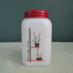 May include: A white spice jar with a red lid. The jar has a rectangular shape with rounded corners. The words "ALL SPICE" are printed vertically on the side, along with a graphic of two potted plants. The jar is likely vintage.