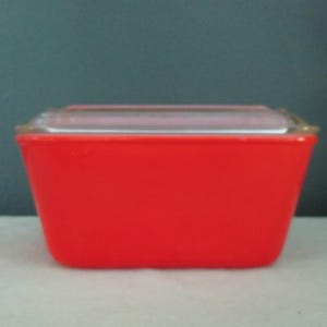 May include: A rectangular, bright red storage container with a clear glass lid. The container has a slightly tapered design, wider at the top and narrower at the base. The lid sits flush on top.