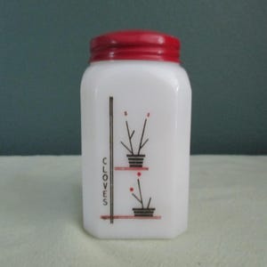 May include: A white glass spice jar with a red lid. The jar features a black and red graphic of two potted plants and the word "CLOVES" in black. The jar has a rectangular shape.