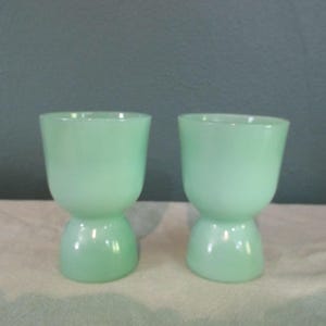 Könnte beinhalten: Zwei hellgrüne Eierbecher aus Glas. Jeder Becher hat einen abgerundeten Boden und eine breitere, zylindrische Oberseite. Die Becher bestehen aus einem durchscheinenden Glasmaterial und stehen auf einer hellen Oberfläche vor einem dunklen Hintergrund.