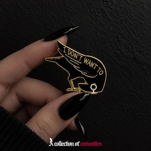 Puede incluir: Un broche de esmalte negro con forma de cuervo y bordes de metal dorado. El ave aparece en una postura hacia abajo con el texto I DON'T WANT TO escrito en letras doradas a lo largo del ala. Un pequeño símbolo dorado se encuentra en la base.
