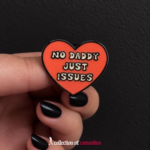 Puede incluir: Un pin de esmalte en forma de corazón con un fondo naranja vibrante y un borde negro fino. El texto NO DADDY JUST ISSUES está impreso en una fuente negra audaz y ondulada en el centro del corazón, creando un diseño gráfico claro y legible.