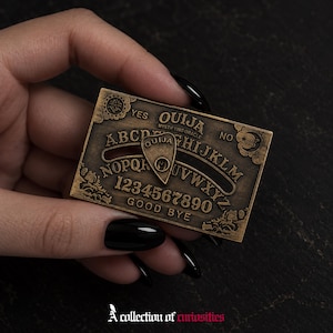 Può includere: Una tavola Ouija rettangolare in metallo color bronzo con l'alfabeto, i numeri da zero a nove, le parole YES, NO, GOOD BYE e OUIJA MYSTIFYING ORACLE. Una planchette triangolare abbinata con la parola OUIJA è posizionata al centro.