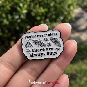 Könnte beinhalten: Eine weiße Emaille-Anstecknadel mit schwarzem, gewelltem Rand, die schwarze Strichzeichnungen verschiedener Insekten sowie den Text "you're never alone there are always bugs" in einer fetten, schwarzen Serifenschrift zeigt.