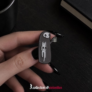May include: A black and grey enamel pin featuring a hooded figure with a skull face holding a small mug in one hand and carrying a white cat with grey spots in the other arm. The design is rendered in a flat, graphic style with clean outlines.