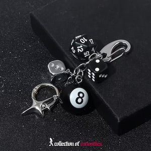 Puede incluir: Un llavero de metal plateado con varios dijes, incluyendo un dado negro de veinte caras con números blancos, un dado negro de seis caras con puntos blancos, un dado plateado, una bola de billar número ocho y un dije de estrella de cuatro puntas.