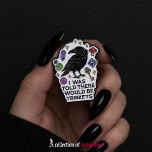 May include: A white enamel pin shaped like a tombstone featuring a black raven, a purple crystal, a green potion bottle, a red mushroom, a blue die, and a gold key. The text reads I WAS TOLD THERE WOULD BE TRINKETS in black capital letters.