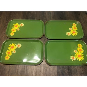 Puede incluir: Un juego de cuatro bandejas de servicio rectangulares de metal verde, cada una con un grupo de flores amarillas y naranjas similares a margaritas en una esquina. Las bandejas tienen esquinas redondeadas y un acabado liso y brillante.