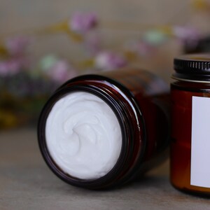 May include: A close-up of a brown glass jar with a white cream inside. The jar is partially open, revealing the creamy texture of the product.