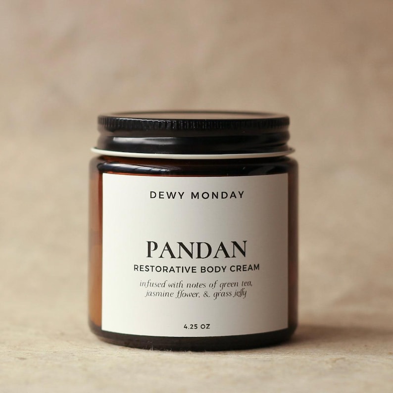 May include: A brown glass jar with a black lid containing Pandan Restorative Body Cream. The label is white with black text that reads "DEWY MONDAY", "PANDAN", "RESTORATIVE BODY CREAM", "infused with notes of green tea, jasmine flower, & grass jelly", and "4.25 OZ".