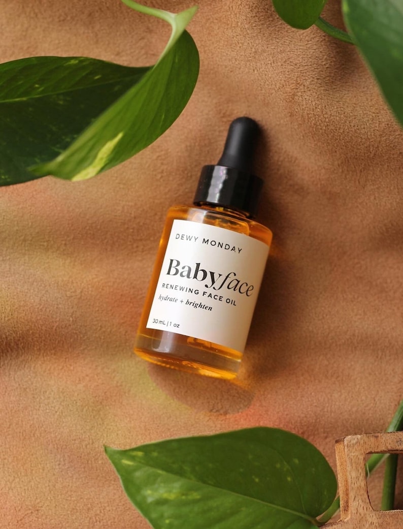 K&ouml;nnte beinhalten: Eine durchsichtige Glasflasche mit schwarzem Tropfer und einem wei&szlig;en Etikett mit der Aufschrift "DEWY MONDAY Babyface RENEWING FACE OIL hydrate + brighten 30 mL | 1 fl oz".