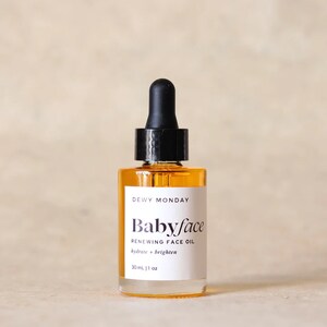 K&ouml;nnte beinhalten: Eine durchsichtige Glasflasche mit einem schwarzen Tropfer oben, gef&uuml;llt mit einer gelben Fl&uuml;ssigkeit. Das Flaschenetikett lautet "DEWY MONDAY Babyface RENEWING FACE OIL hydrate + brighten 30mL | 1 oz"