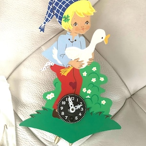 May include: A vintage wooden clock featuring a child holding a white goose. The child wears a blue cap, shirt, and red pants. The clock face is black with white numbers and hands, set on a green base with white flowers.