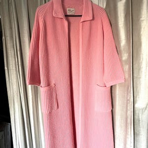 May include: A light pink, ribbed-knit cardigan with a collar and two front pockets. The longline sweater has short sleeves and hangs on a black hanger. The label on the inside of the collar is visible.