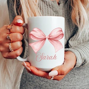 Pode incluir: Caneca de cerâmica branca com um laço rosa e o nome "Sarah" impresso nela.