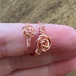 May include: Two copper wire rings, each featuring a coiled rose-shaped design. The wire is wrapped to form a central spiral flower motif with a smooth band. The metal has a warm, metallic copper tone and a polished finish, showcasing intricate wirework.