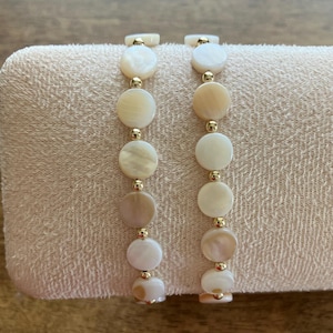 May include: Two bracelets featuring alternating round, flat, off-white mother-of-pearl shell discs and small, polished gold-tone metal beads. The circular shell components display natural variations in colour and lustre, arranged in a single strand.