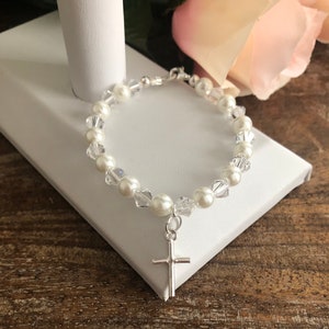 Puede incluir: Un brazalete de perlas blancas y cristal con un dije de cruz de plata. El brazalete está sobre una superficie blanca con una rosa rosa en el fondo.