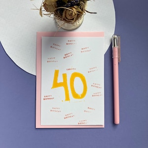Peut inclure: Carte d'anniversaire avec le chiffre 40 en jaune, entouré du texte "Happy Birthday". La carte est sur un fond rose, avec un stylo rose et un petit arrangement de fleurs séchées.