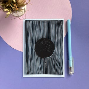Op de afbeelding: Een zwart-wit kunstafdruk met een zwarte cirkel op een achtergrond van verticale lijnen. De afdruk staat op een witte kaart, met een lichtblauwe pen en een kleine vaas met gedroogde bloemen. De achtergrond is roze en paars.