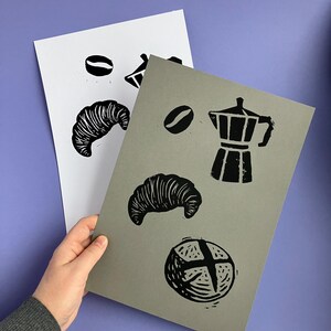 Op de afbeelding: Twee prints met zwarte linosnede illustraties van koffiebonen, een croissant, een koffiepot en een rond brood. Eén print is op wit papier, de andere op grijs papier. De prints worden door een hand tegen een paarse achtergrond gehouden.