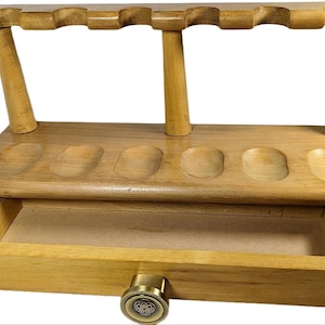 May include: Wooden pipe rack with six slots for pipes and a drawer. The rack is made of light brown wood and has a brass knob on the drawer.